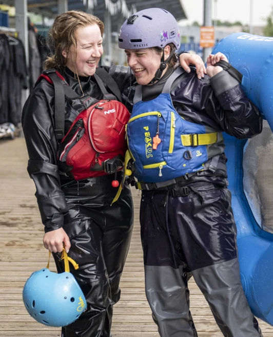 Two smiling students in waterproof gear share a moment after a water sports session, one holding a raft. Two smiling students in waterproof gear share a moment after a water sports session, one holding a raft.